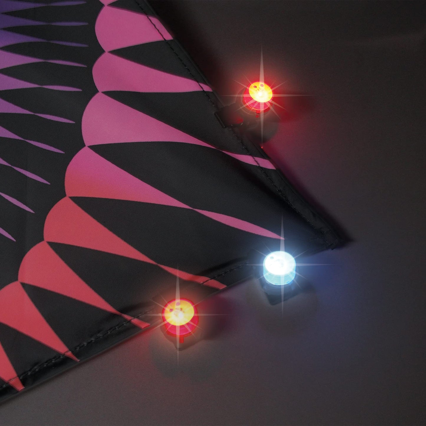 A photo of a kite on a white background in the dark with three WindNSun UFO Mini Lights flashing red and blue cliped along the edge