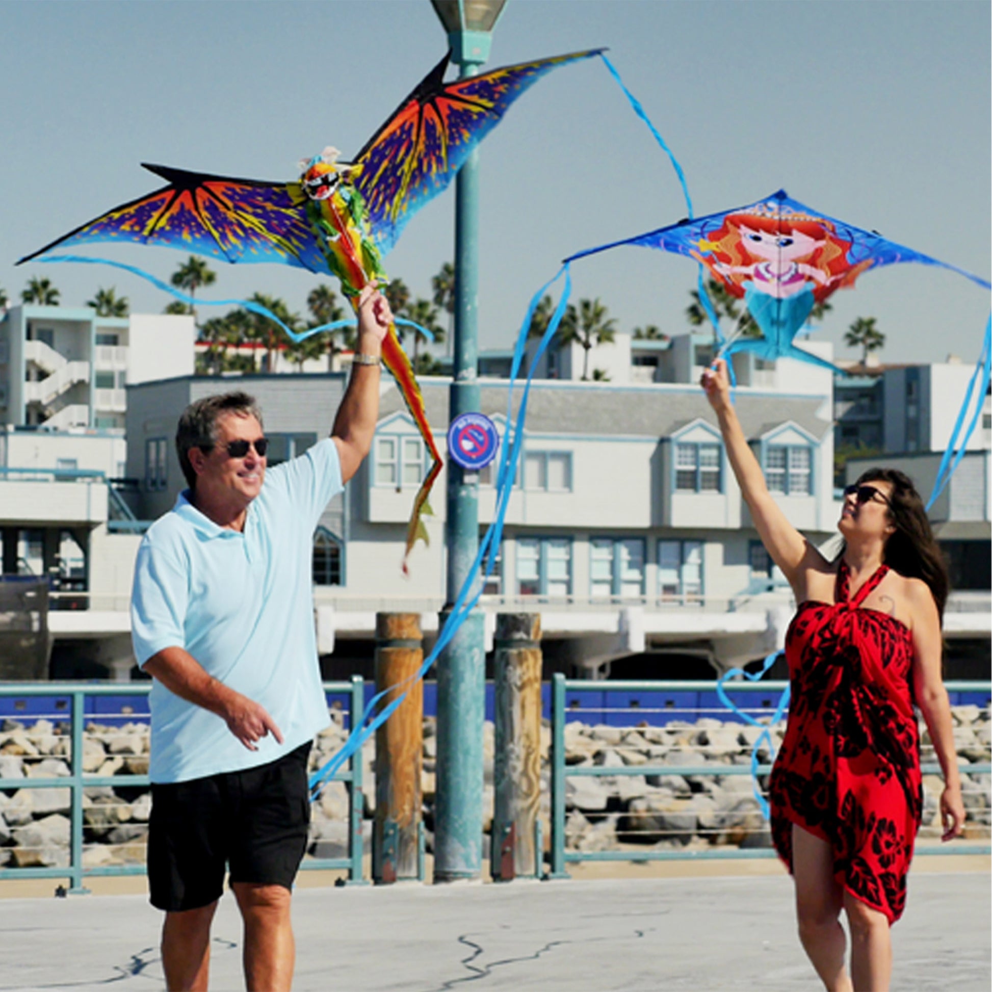 A man and woman walking along a beach boarwalk, each holding a kite over their head