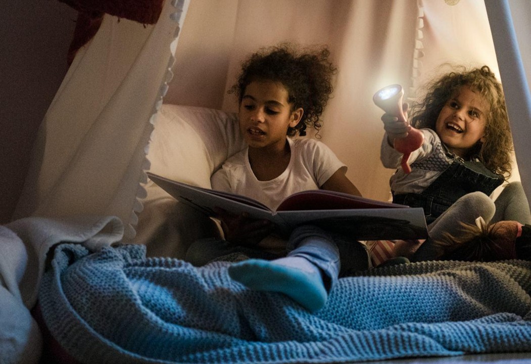 Two children reading together in a blanket fort with a handheld light