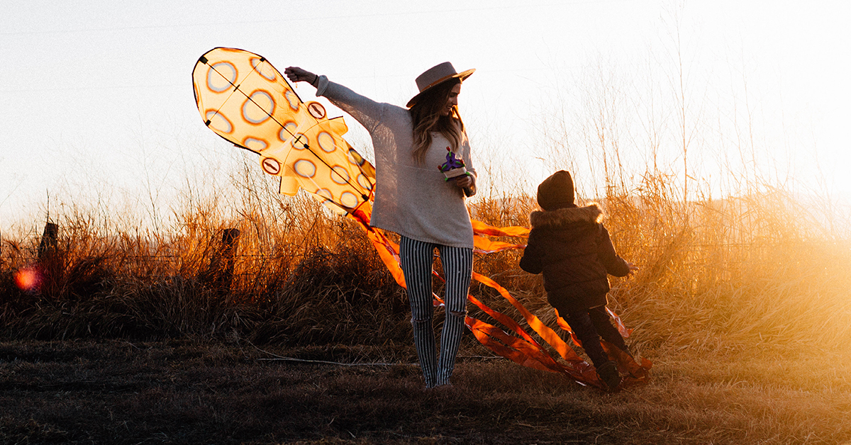 A parent and child flying a bright orange kite outdoors at sunset